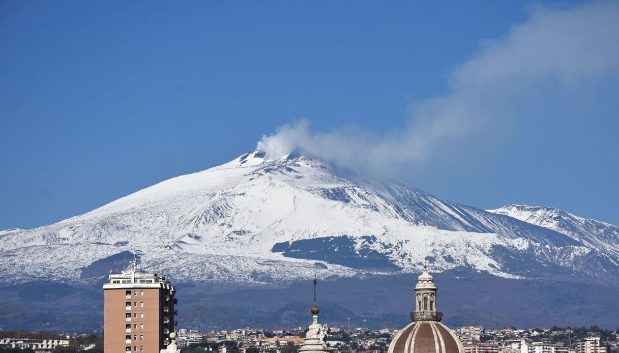 Panoramic view of snow-capped Mount Etna volcano emitting smoke above the skyline of Catania, Sicily.