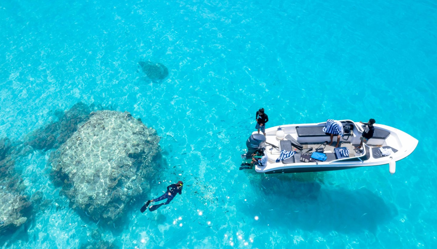 Aerial view of a boat and snorkeler in crystal clear turquoise tropical water near a coral reef.