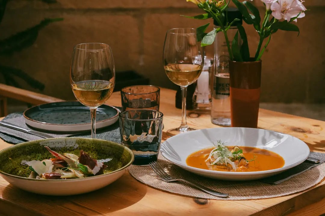 Mesa montada con comida encima, se ve deliciosa, alcachofas y sopa de pescado. Hay flores bonitas en el restaurante Es Mestra