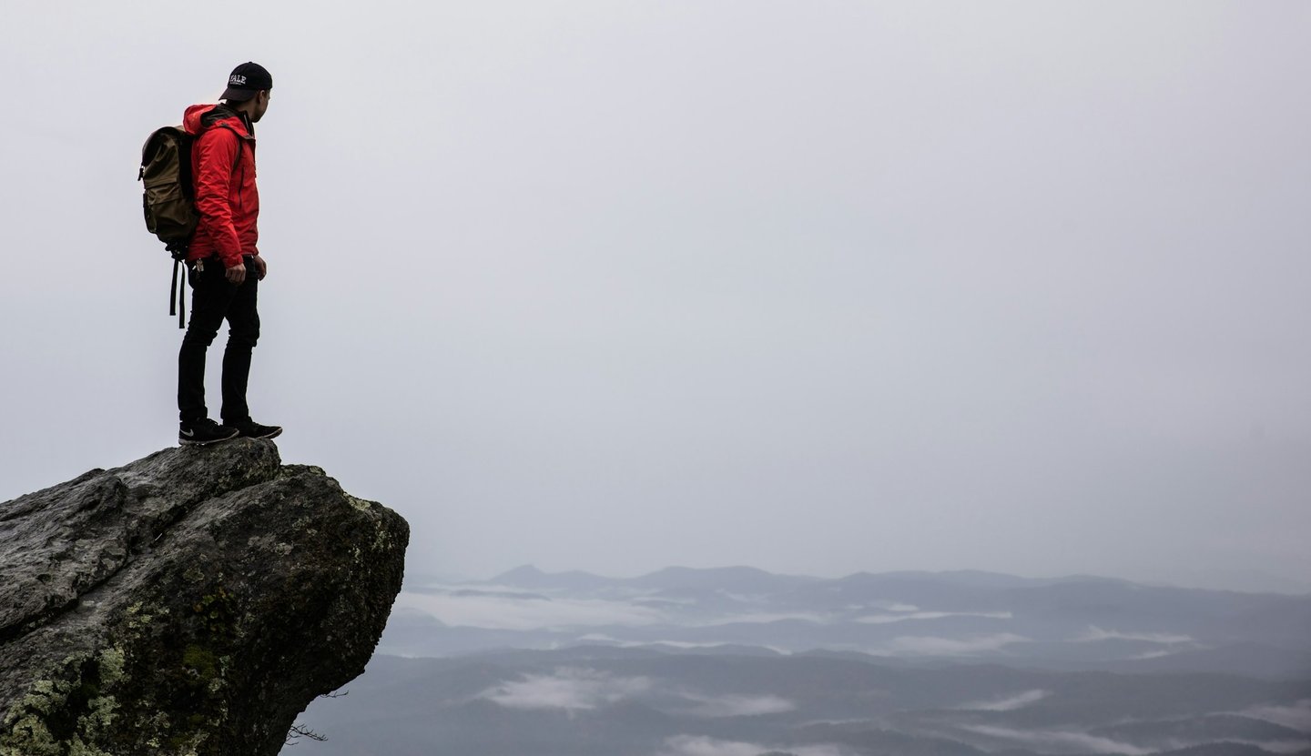 A man in a red jacket standing on a cliff rock, looking out at the plains against a cloudy grey sky