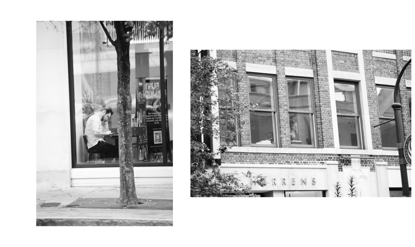 Black and white street photography of a person sitting in a cafe window next to a brick building.