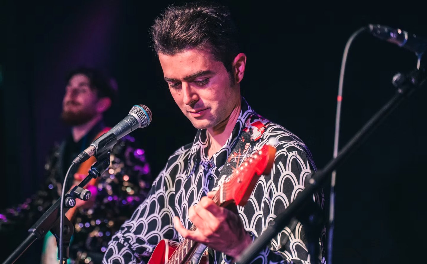 Olly Williams playing a red guitar and singing into a microphone on stage in Brighton.