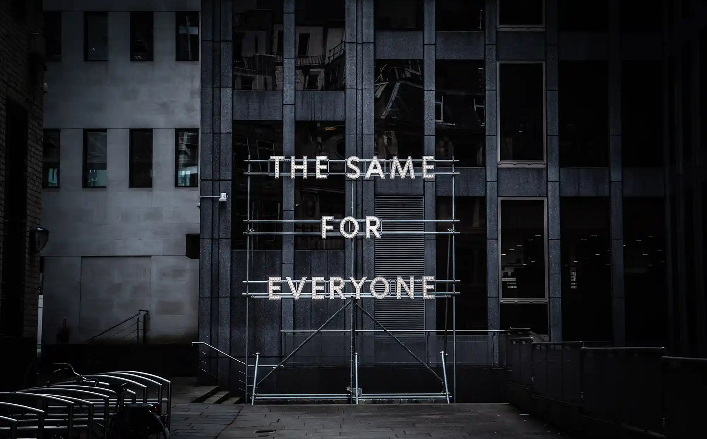 Illuminated neon sign saying The Same For Everyone on metal scaffolding in front of a modern office building.