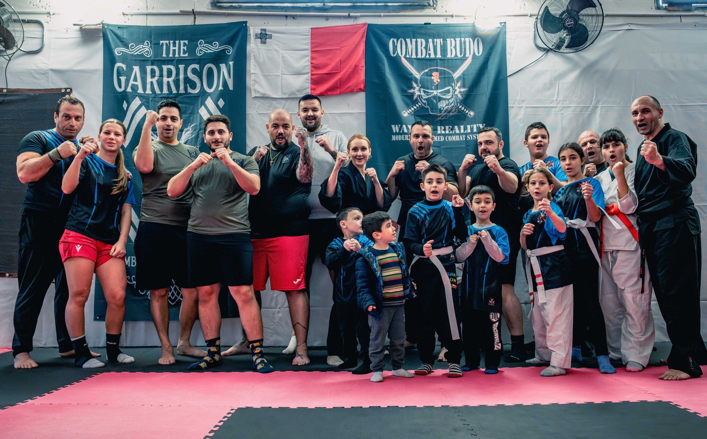 Adults and children group posing in a Combat Budo martial arts dojo for self-defense training.
