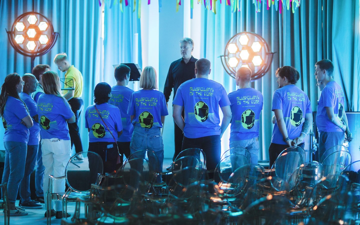 A group of employees in blue corporate t-shirts attending a stage presentation at a tech event.