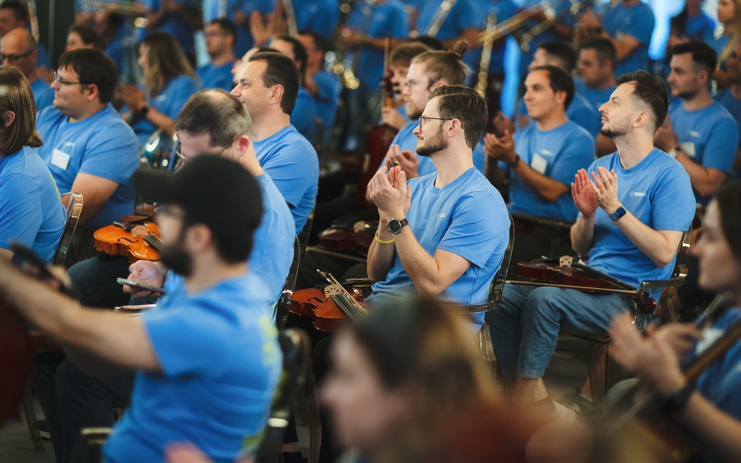 Musicians in blue t-shirts clapping during an orchestral rehearsal with violins.