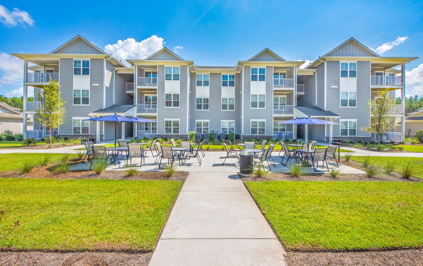 Ground-level view of outdoor patio seating with umbrellas between apartment buildings at Arrogate Village in Summerville, SC