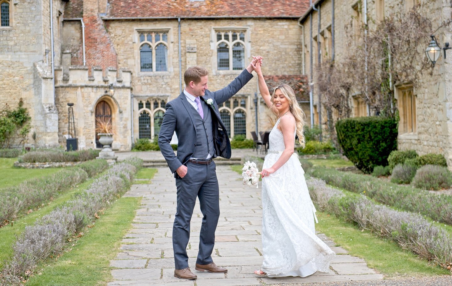 bride in white dress, groom in navy suit, dancing in front of stately home, groom whirling bride, both happy