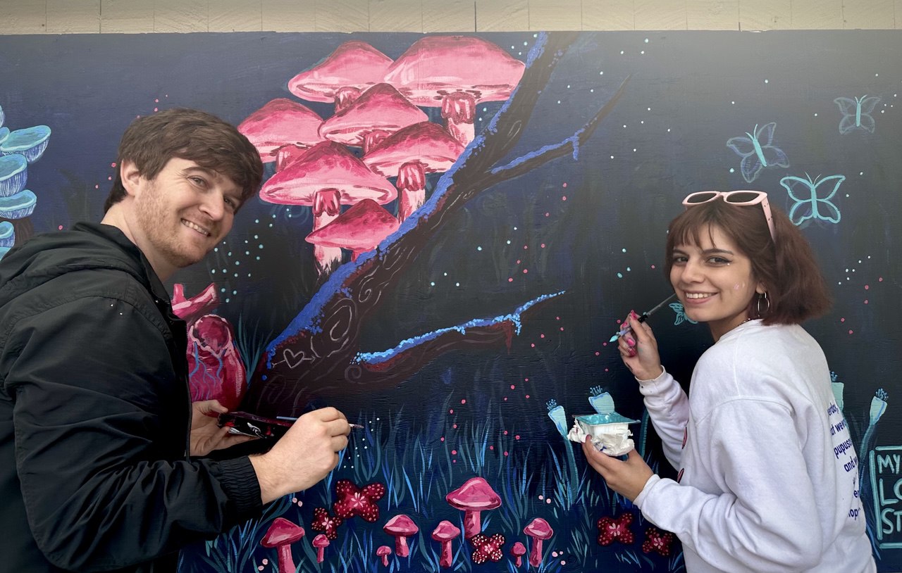 Two artists painting a whimsical mural featuring glowing pink mushrooms and butterflies.