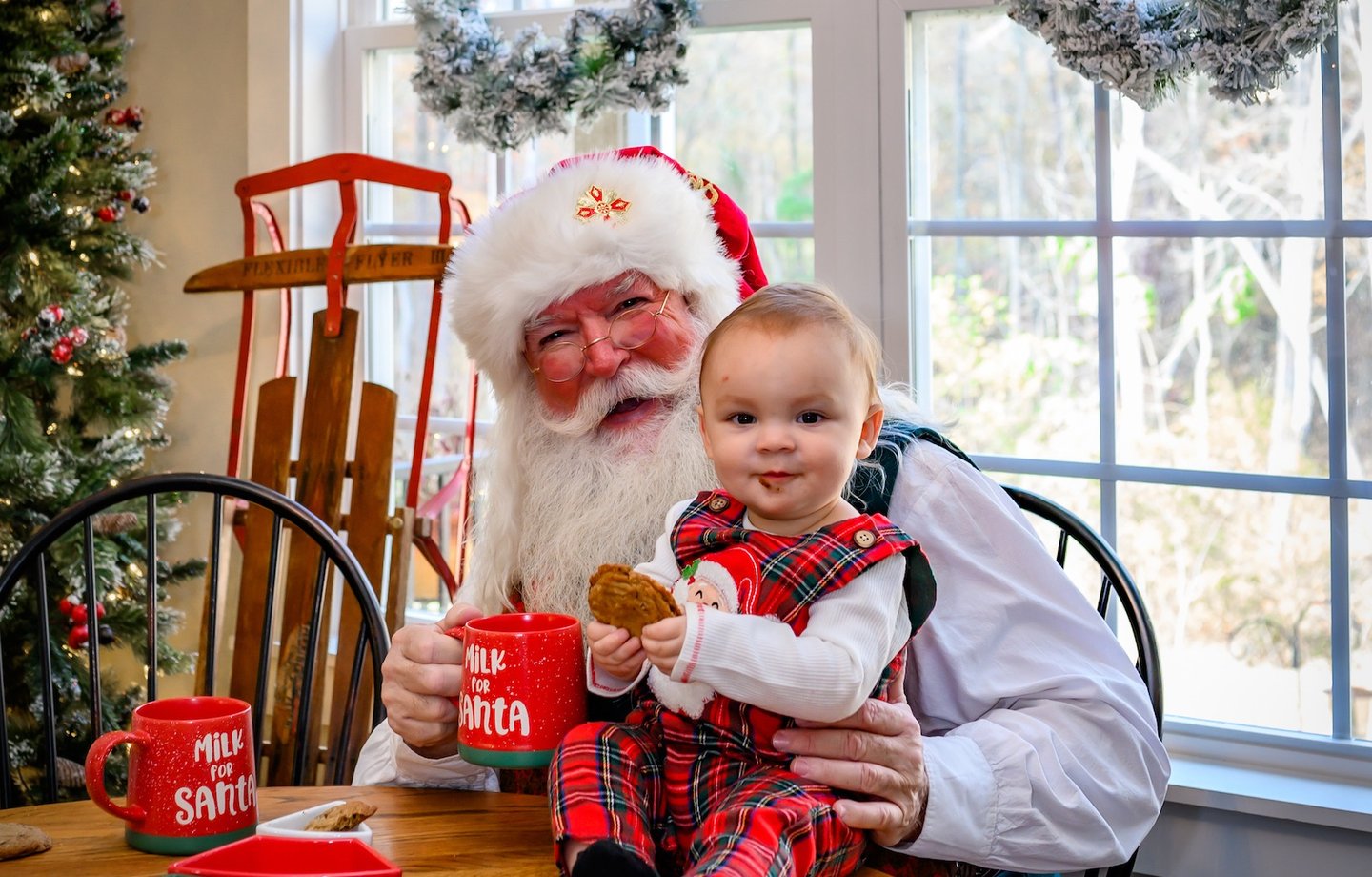 Santa Claus sits at a wooden table with a baby in plaid pajamas, enjoying cookies and mugs of milk.