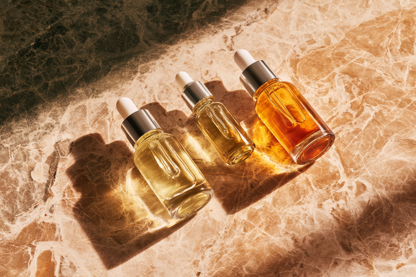 Three glass dropper bottles of facial oil and serum on a luxury marble surface with sunlight shadows.