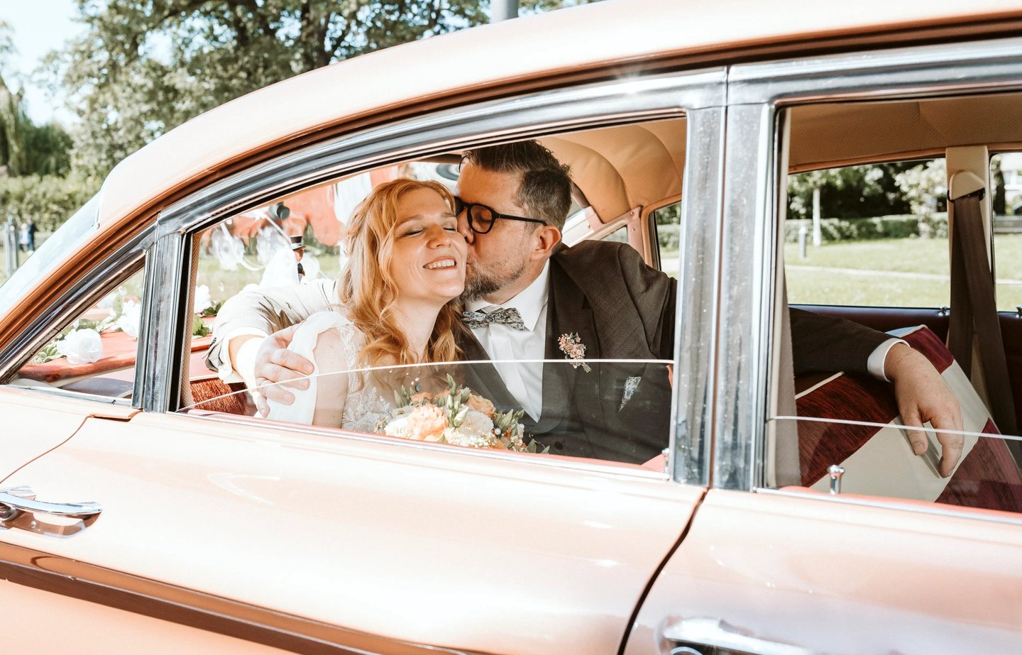 Braut und Bräutigam sitzen glücklich in einem Oldtimer, der Bräutigam küsst die Braut liebevoll auf die Wange, Brautstrauß im