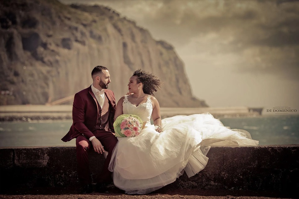 Mariés assis sur muret face à la mer et falaise, costume bordeaux et robe princesse, bouquet pastel.