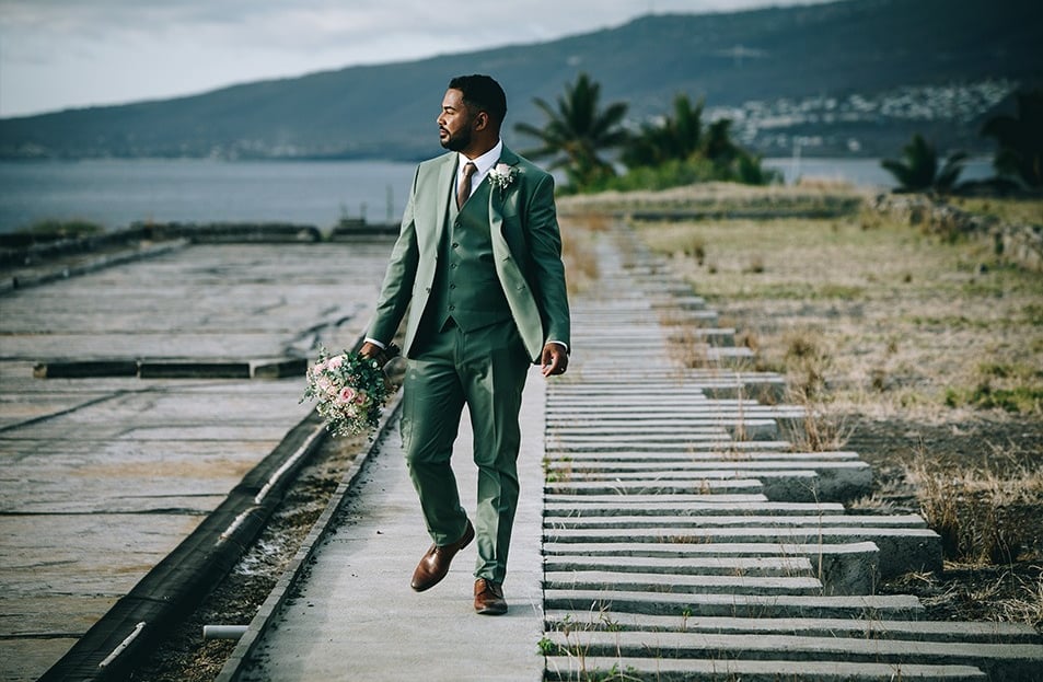 Marié élégant en costume trois pièces vert sauge tenant le bouquet de mariée, marche sur un ponton.
