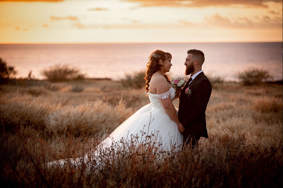 Mariés se regardant au coucher de soleil, robe épaules dénudées et costume noir, face à la mer. mer.