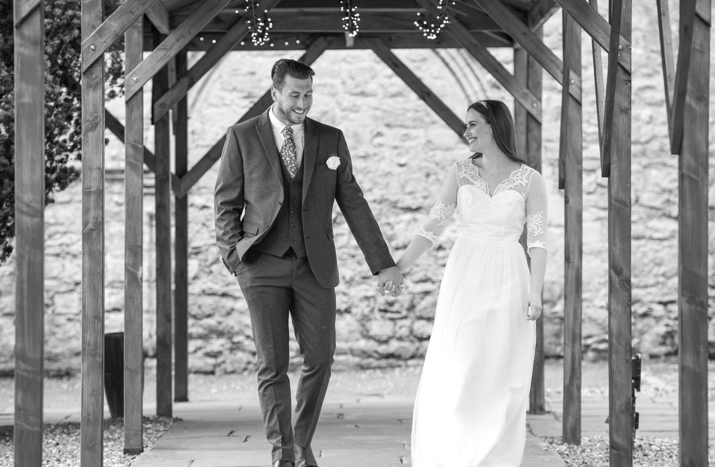 black & white photo of bride and groom walking and holding hands, both smiling, wooden canopy is framing them