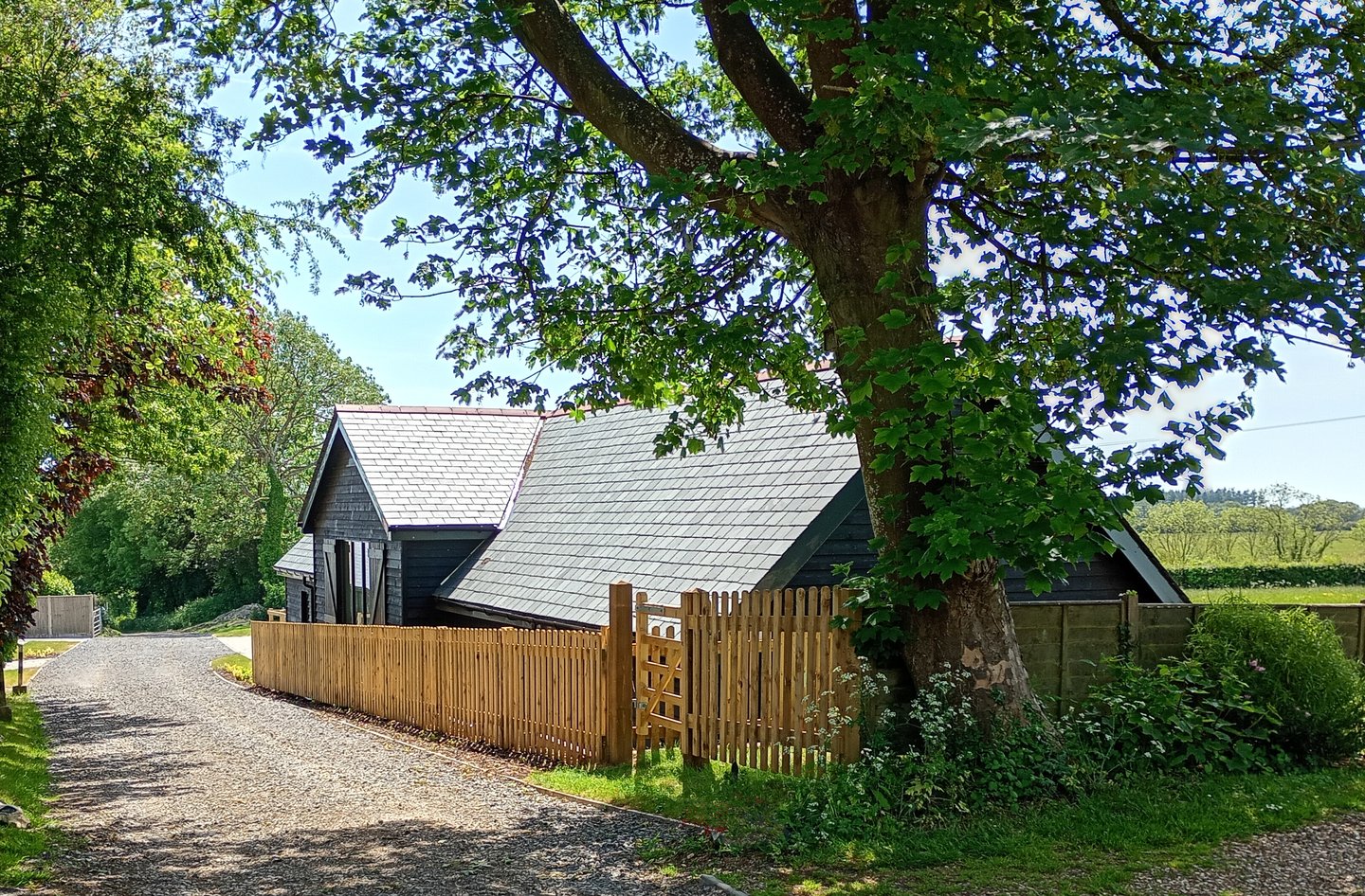 A black timber barn conversion with a slate roof behind a wooden picket fence and large tree.