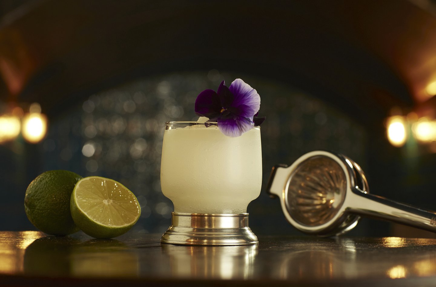 White cocktail with a pansy flower garnish next to cut limes and squeezer in an art deco bar