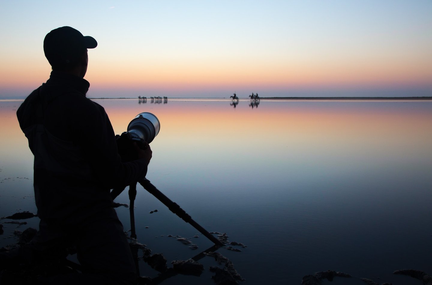 A wildlife photographer silhouettes a sunset over a calm lake with horses in the distance.