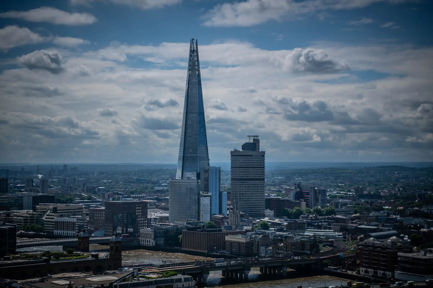 Aerial view of The Shard skyscraper towering over the London city skyline under a cloudy blue sky.