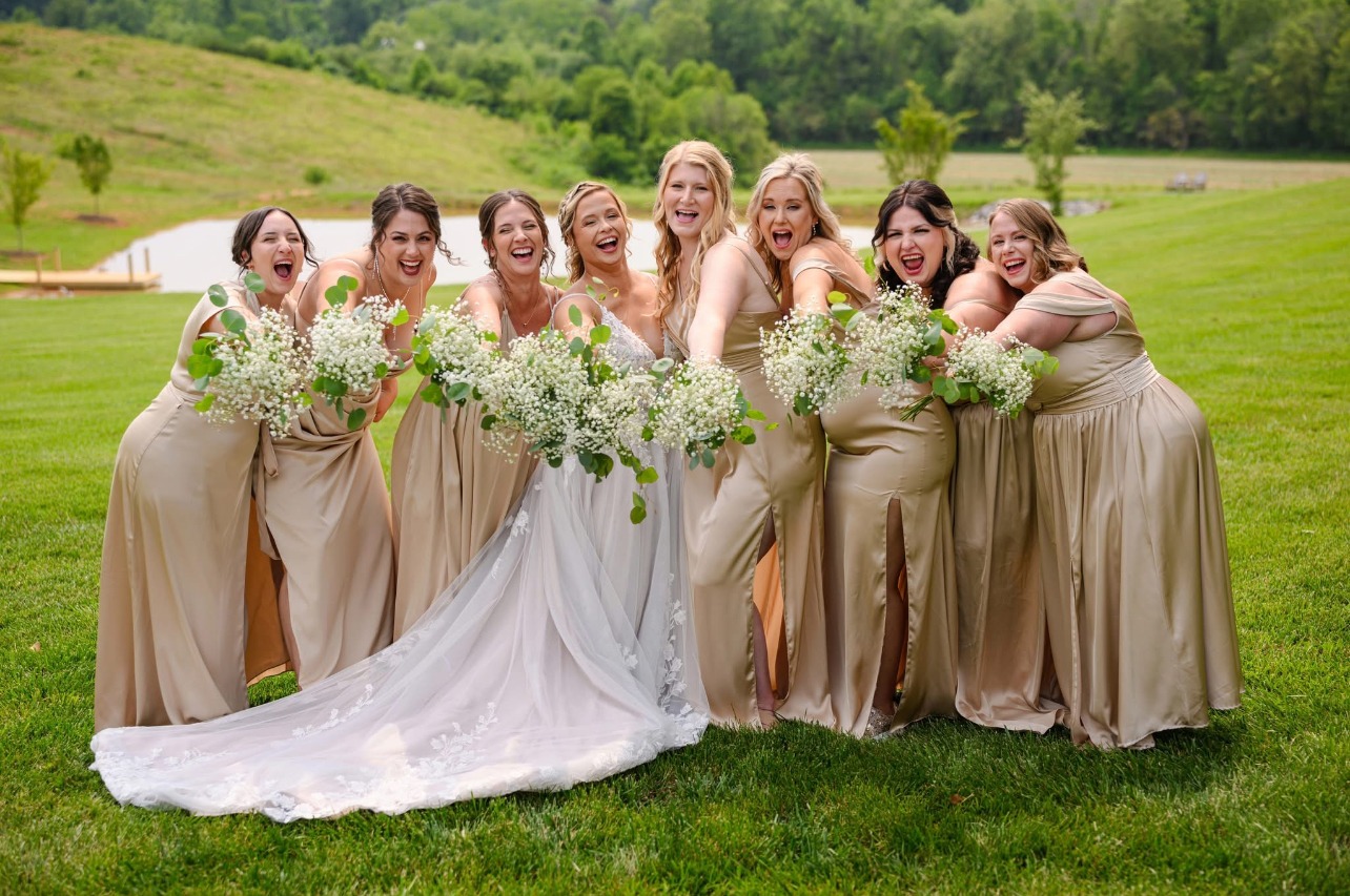 Bride surrounded by bridesmaids in gold dresses holding white bouquets, smiling outdoors, featuring wedding hair and makeup.