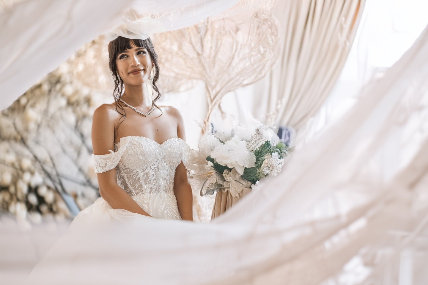 Ritratto emozionale di una sposa sorridente, avvolta da veli e luce soffusa, con bouquet chiaro.