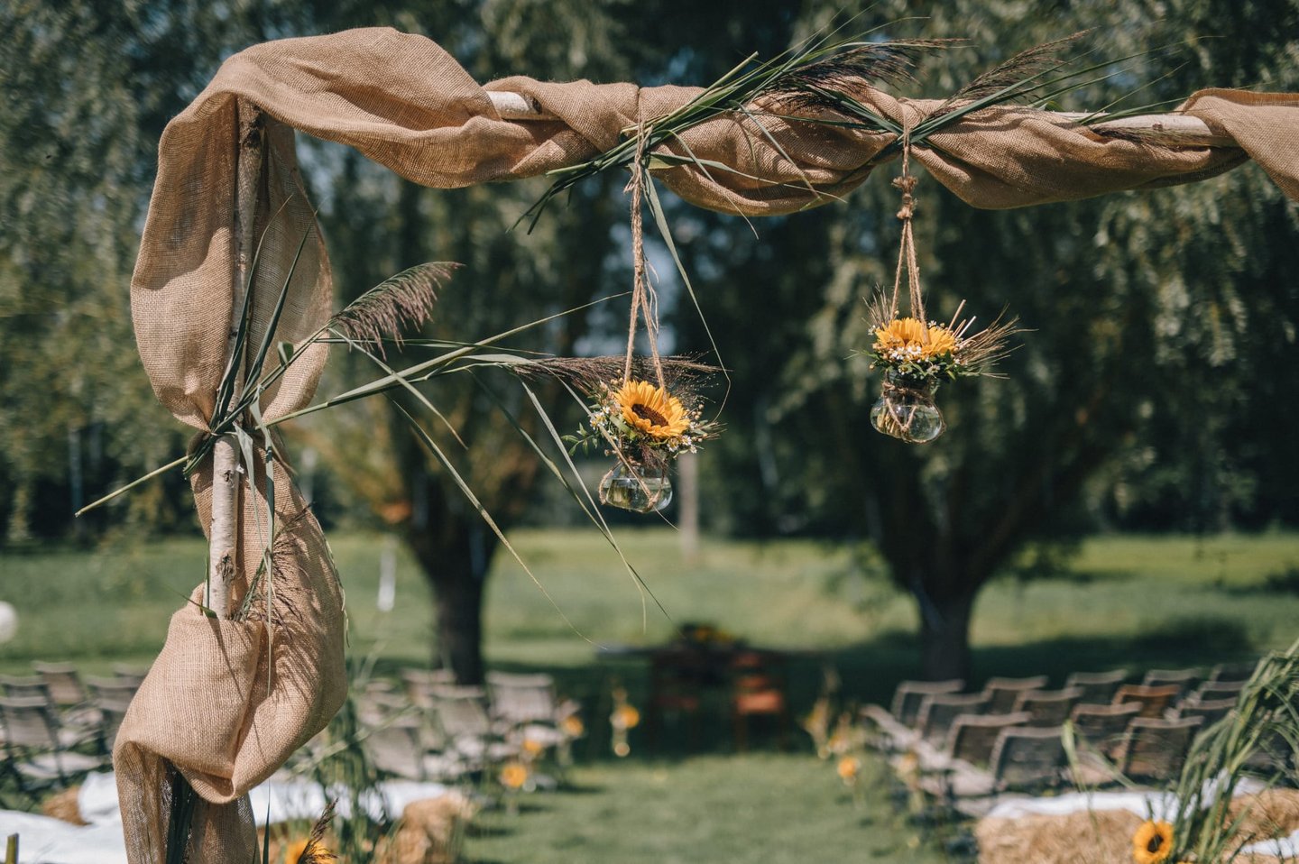 Rustikaler Traubogen im Freien, dekoriert mit Jute-Stoff und hängenden Sonnenblumenvasen 