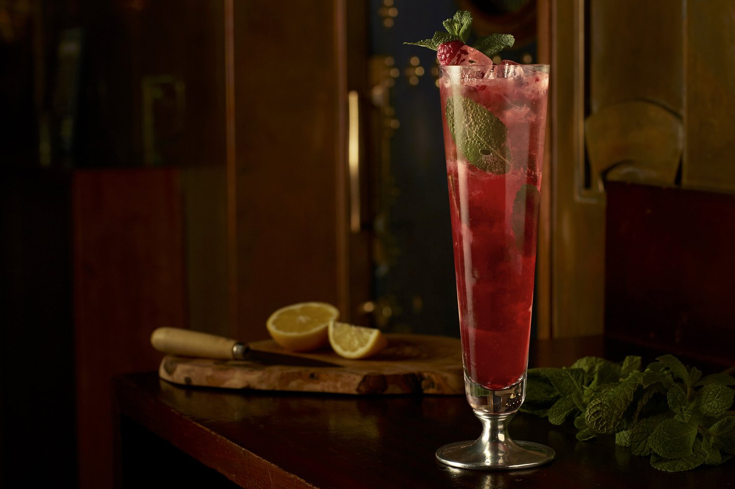 Red cocktail in a tall glass on a wooden table with mint and cut lemons