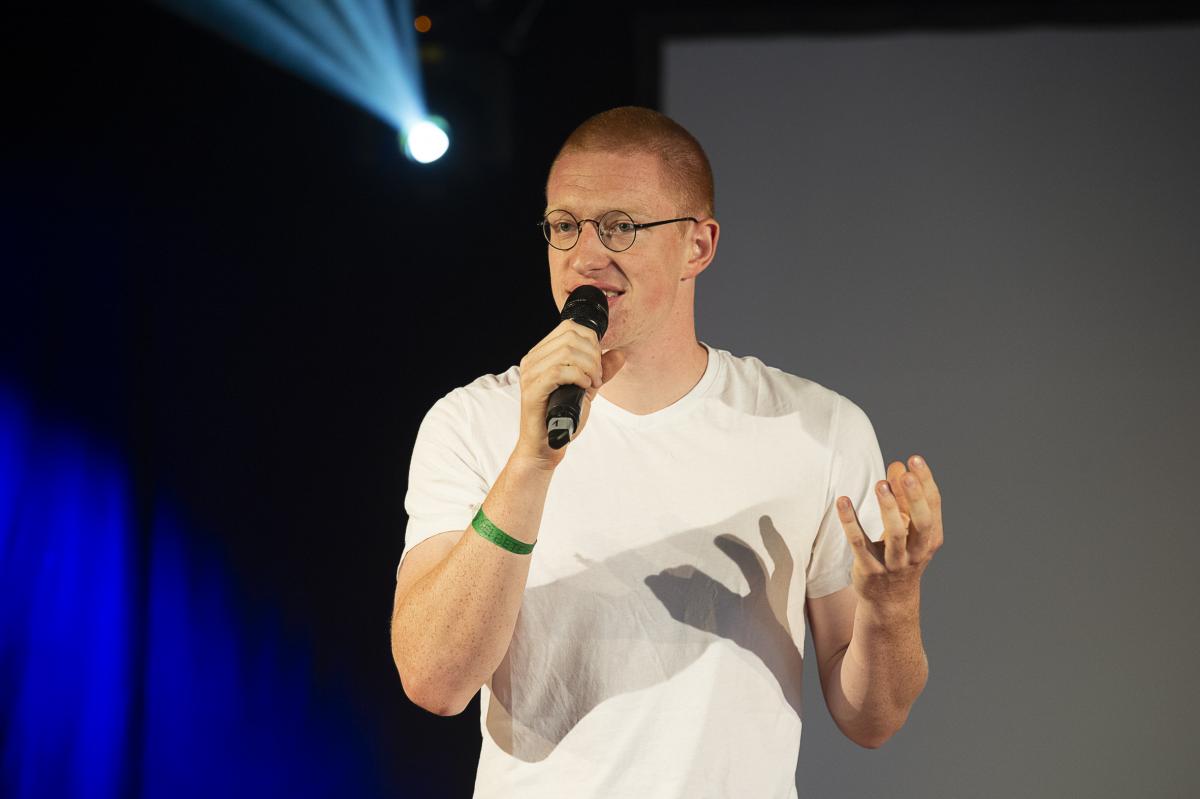 Przemysław Żuk, kurz Przemek, steht auf einer Bühne und spricht in ein Mikrofon. Er gestikuliert. Im Hintergrund Bühnenlicht.