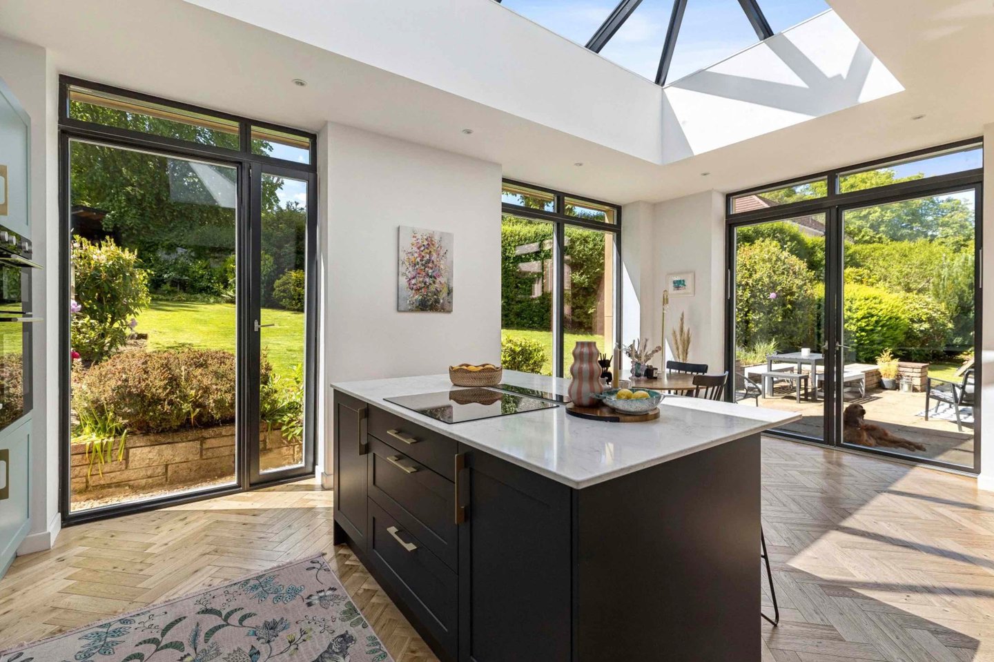 Modern open-plan kitchen featuring a dark island, skylight, and large glass doors opening to a lush garden.