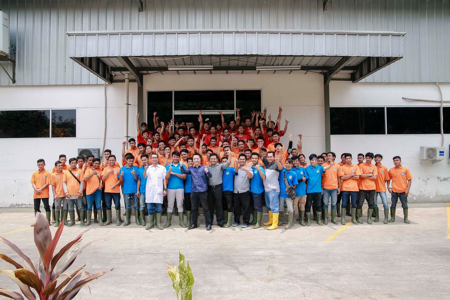 A large group of industrial warehouse employees in orange and blue uniforms posing outside a factory building.