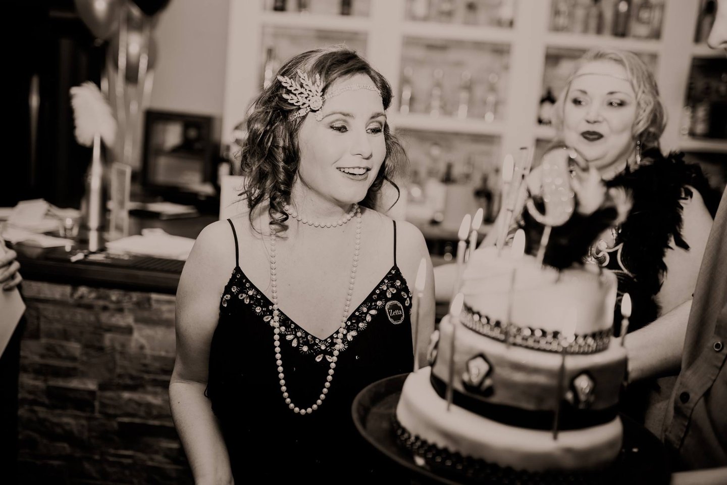 woman in 1920s outfit smiling at a birthday cake. another woman is smiling at the birthday girl.