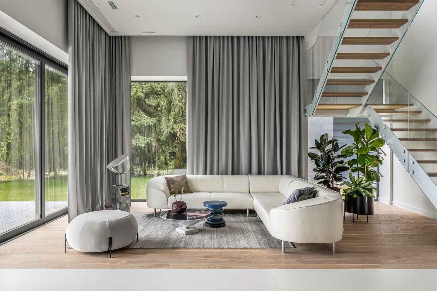 Przestronny salon z białym narożnikiem, szklane schody i widok na las przez wysokie okna.