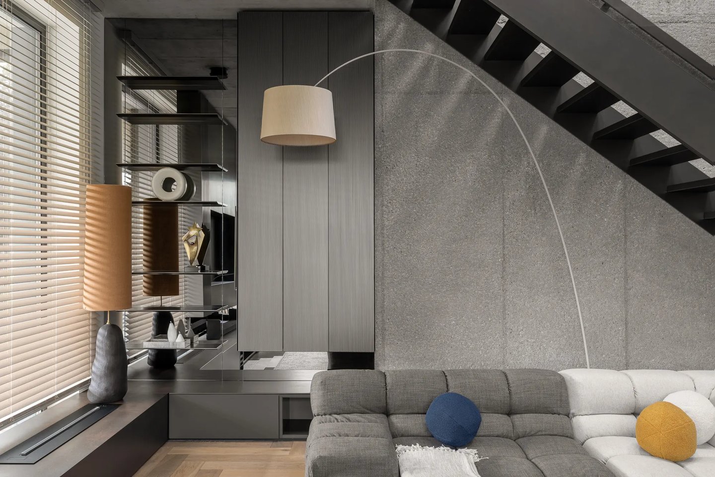 Kącik wypoczynkowy w salonie, szklany regał, lampa łukowa i ściana z architektonicznego betonu.