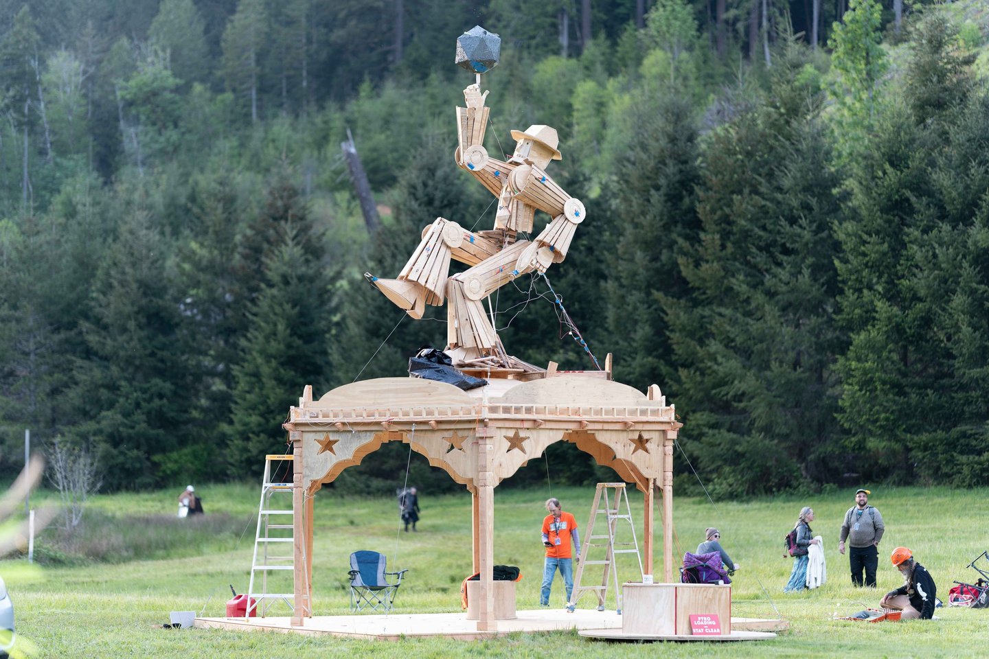 A large wooden figure sculpture atop a gazebo in a forest clearing for an outdoor art installation.