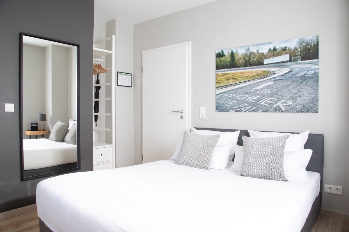 Modern minimalist bedroom with a white double bed, full-length mirror, and racetrack wall art.