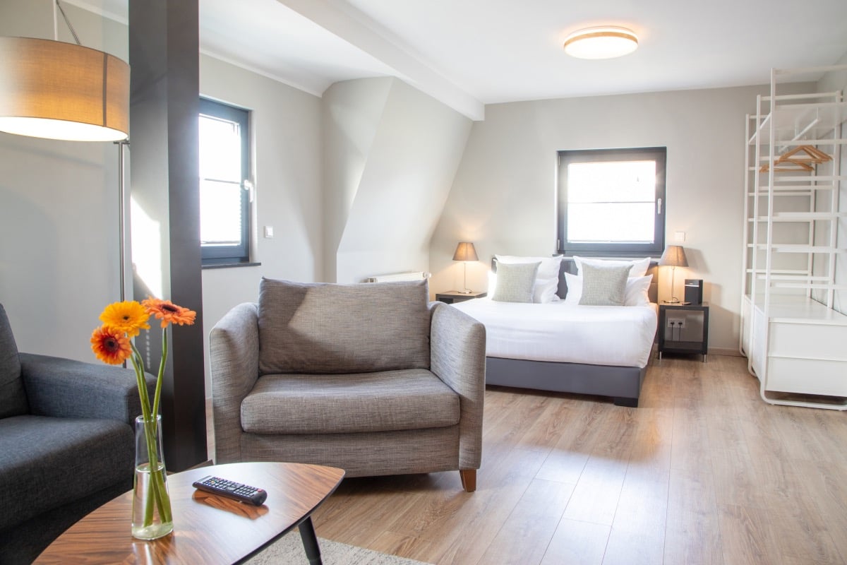 Modern hotel studio apartment with grey armchairs, king size bed, and bright hardwood floors.