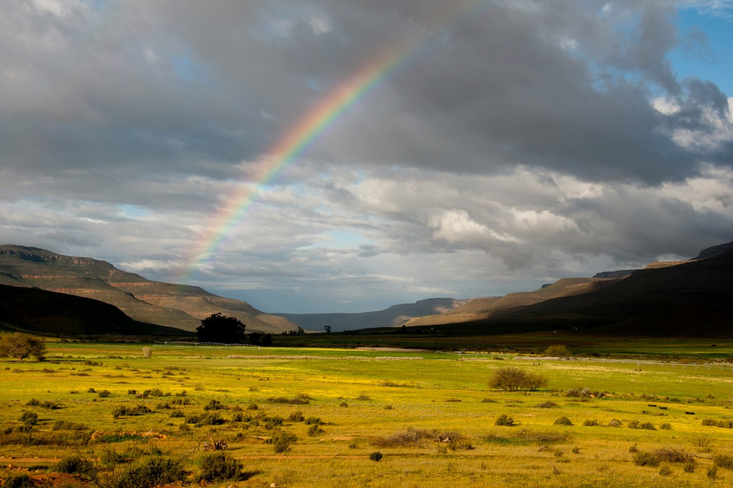 Rainbow across a cloudy sky with some fields in the foreground and a circular mountain range as backdrop