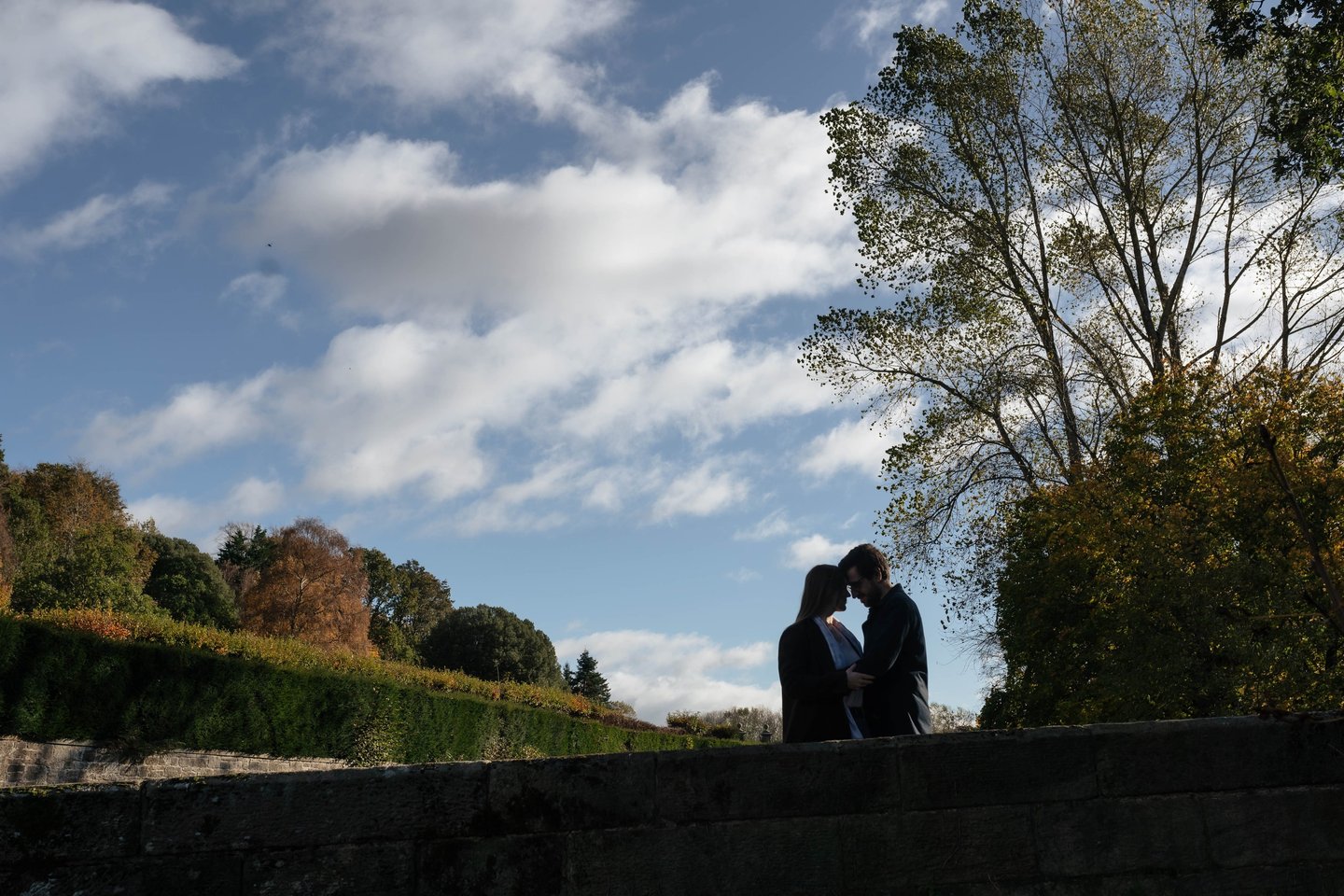 Silhouette of a romantic couple embracing on a stone wall in a scenic autumn park under a blue sky.