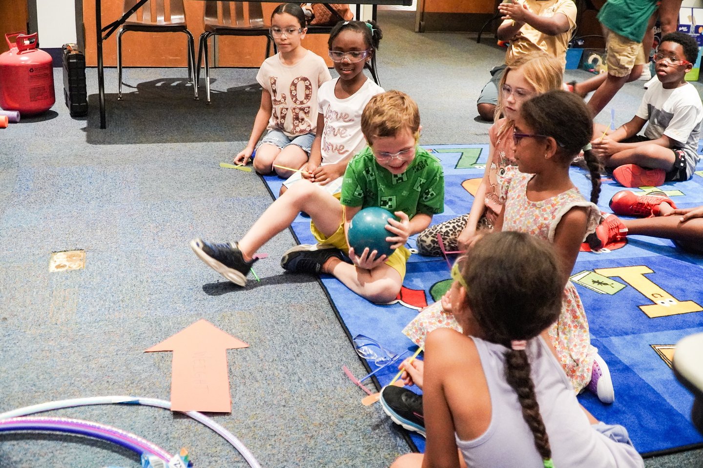 a group of children sitting on a rug in a classroom passing a weighted ball STEM