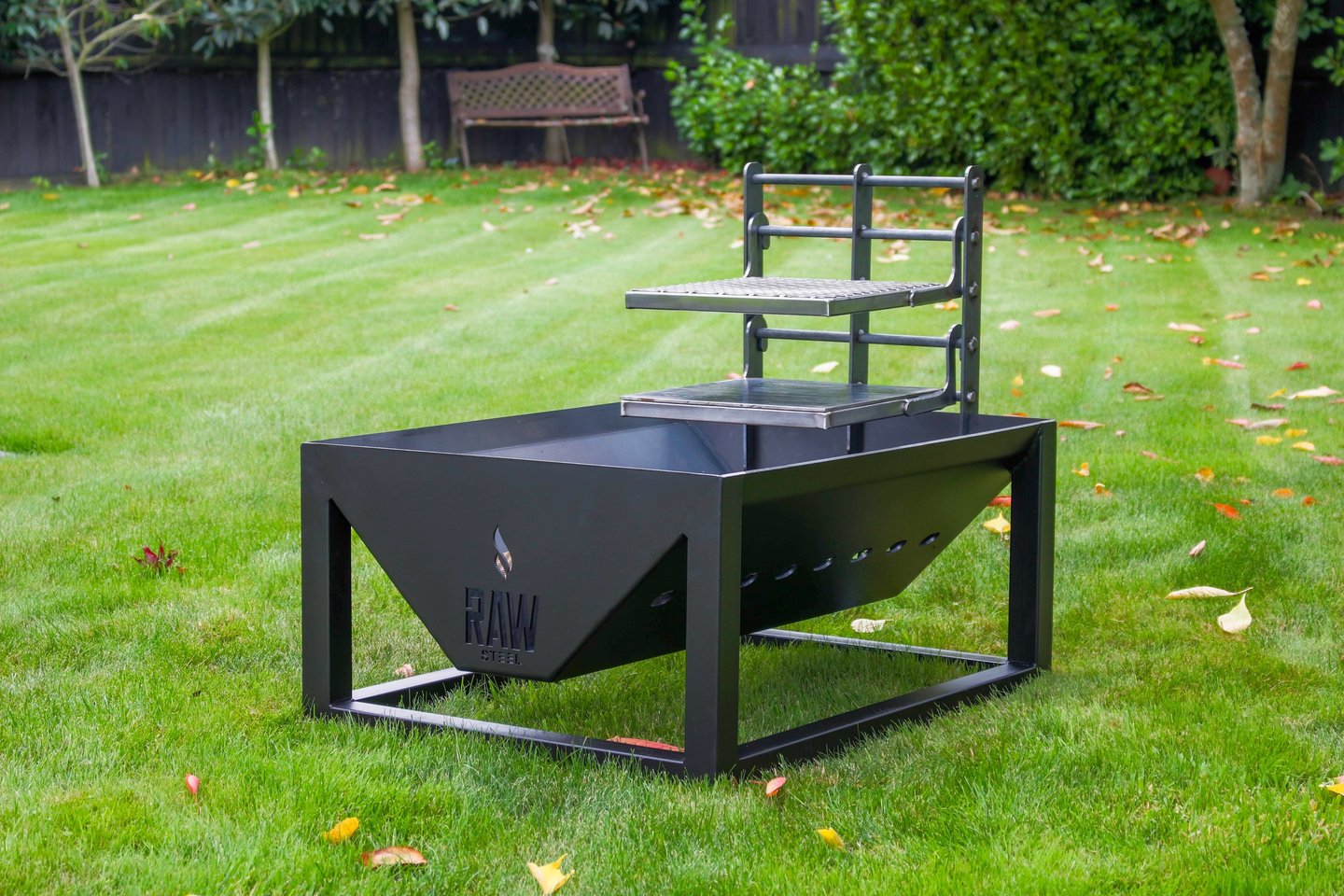A black Raw Steel fire pit with an adjustable grill grate on a green backyard lawn.