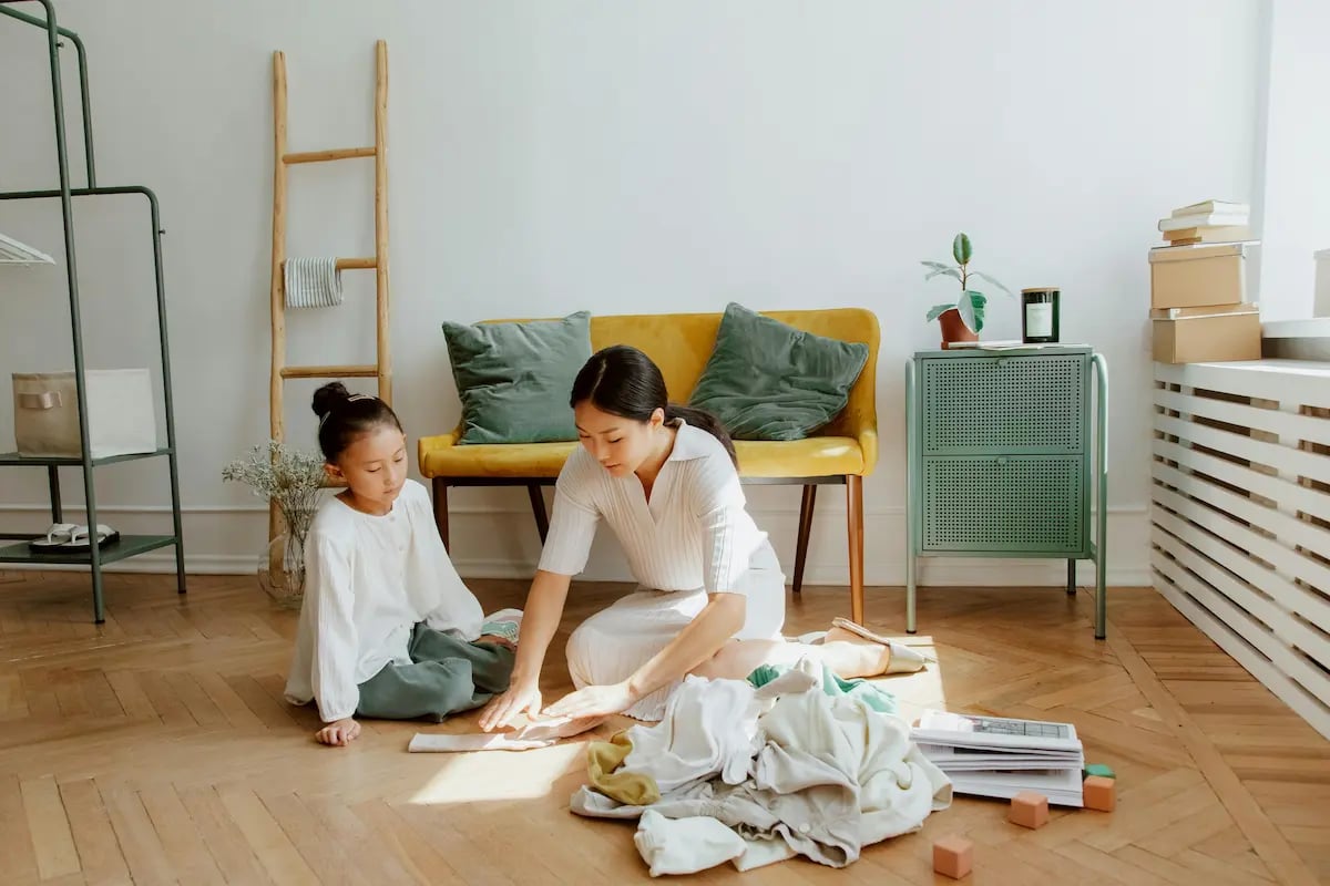 A mother and daughter organizing clothes on a wooden floor in a modern, sunlit living room.