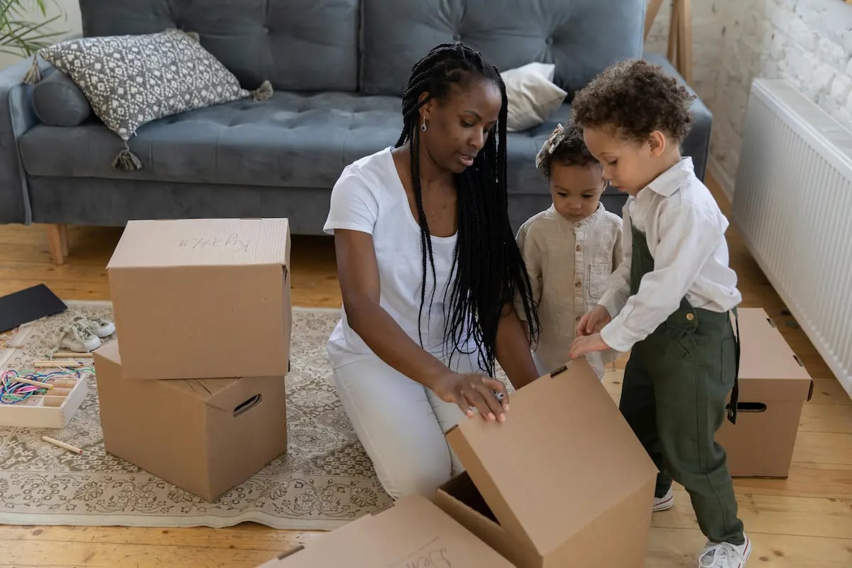 A mother and two young children unpacking cardboard boxes in their new living room.