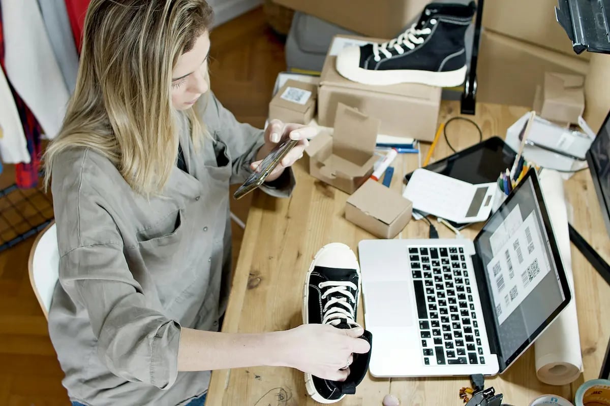 A small business owner photographs shoes on a wooden desk with a laptop and packaging, managing her online store.