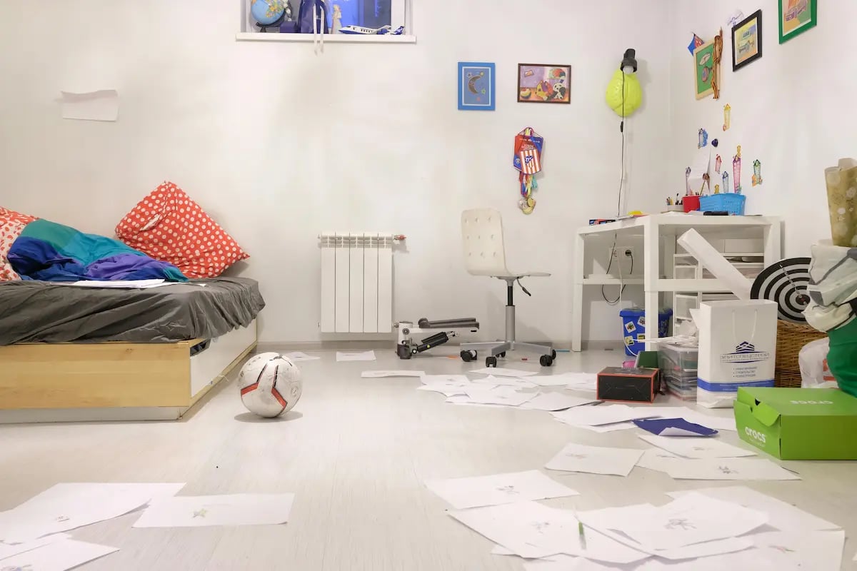 Messy child's bedroom with white walls, scattered drawings on the floor, and a soccer ball.