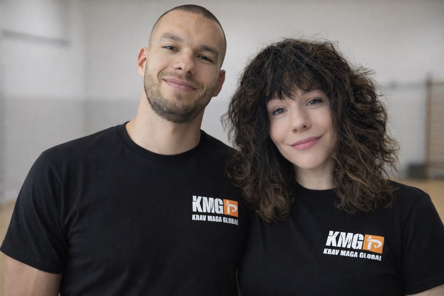 Smiling male and female Krav Maga Global instructors wearing black KMG t-shirts in a gym.