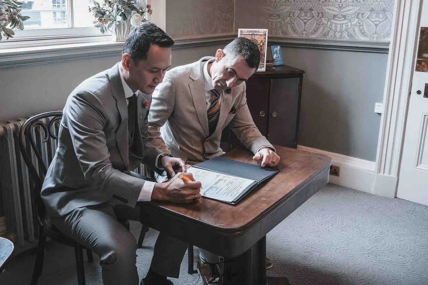 A gay couple in elegant suits signing their marriage certificate during a wedding ceremony.