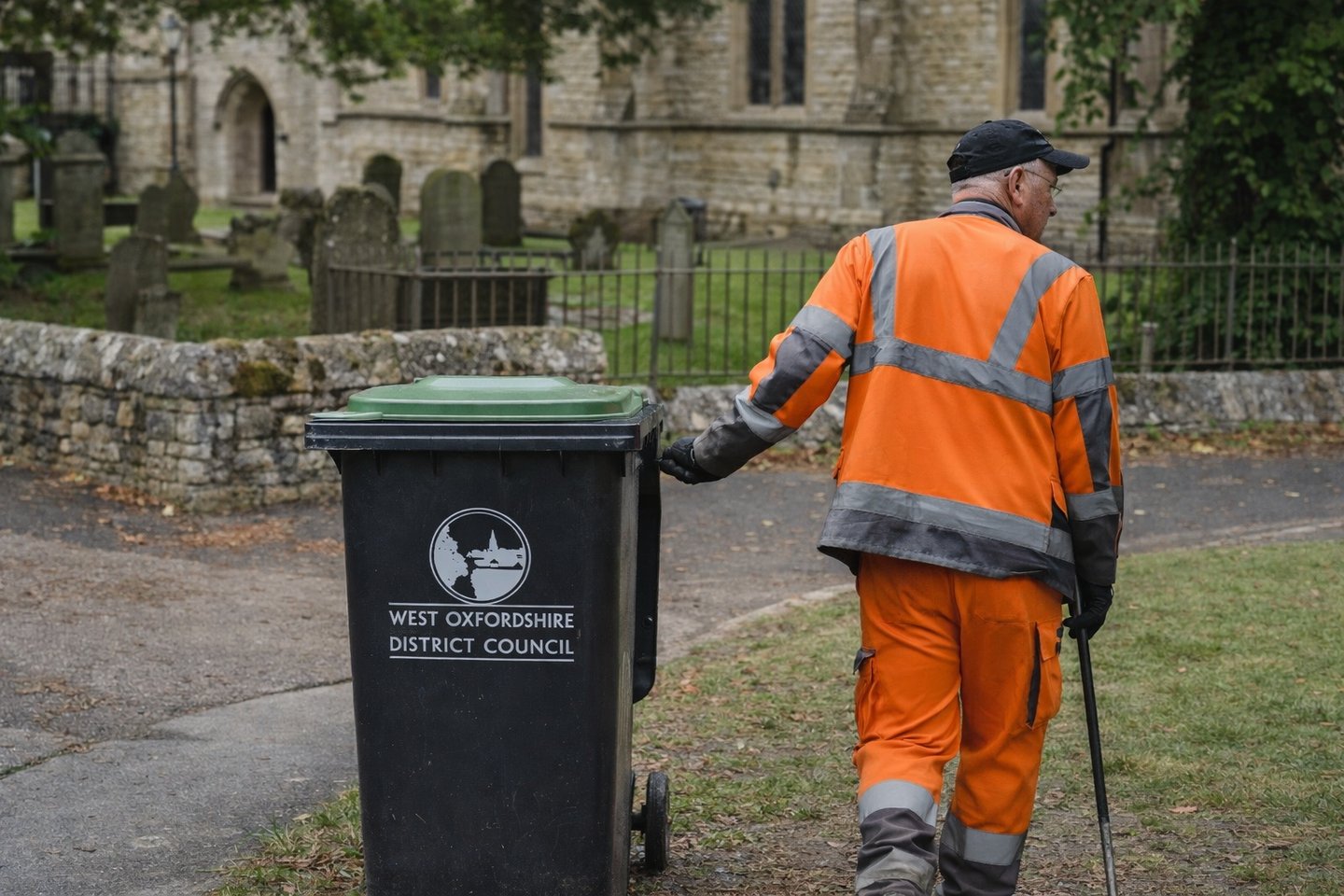 A West Oxfordshire District Council worker in high-visibility orange collects a wheelie bin near a church