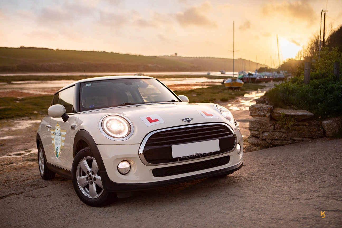 White Mini Cooper driving school car with L-plates parked at a coastal harbor at sunset.