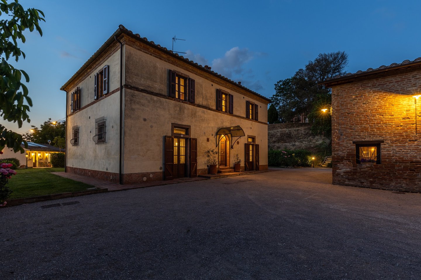 A historic Tuscan villa exterior at dusk with warm glowing lights and a gravel courtyard.